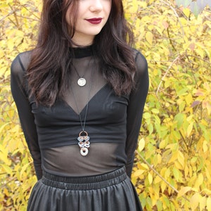 May include: A woman with long dark hair is wearing a black sheer top with a silver and copper necklace. She is also wearing a black skirt. The background is a blurry image of yellow and green leaves.