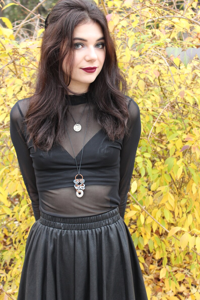 May include: A woman with long dark hair is wearing a black sheer top with a silver and copper necklace. She is also wearing a black skirt. The background is a blurry image of yellow and green leaves.