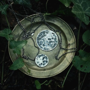 May include: Two silver necklaces with hammered round pendants on a green leafy background. The necklaces are on a brass plate.
