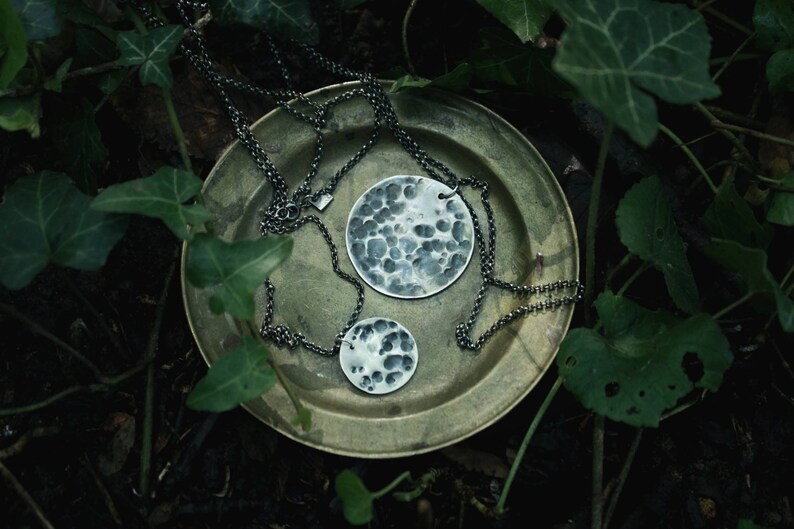 May include: Two silver necklaces with hammered round pendants on a green leafy background. The necklaces are on a brass plate.