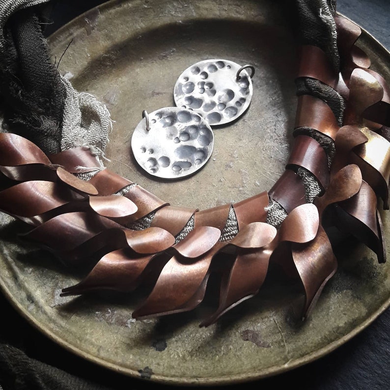May include: A copper and silver necklace with a textured, hammered metal design. The necklace is made of overlapping, leaf-shaped pieces of copper and features two silver pendants with a textured, hammered surface.