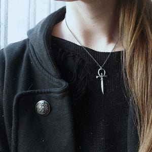 May include: A person wearing a black hooded jacket with a silver dagger pendant necklace. The pendant is a stylized dagger with a circle at the top. The person is wearing a black and white striped shirt underneath the jacket.