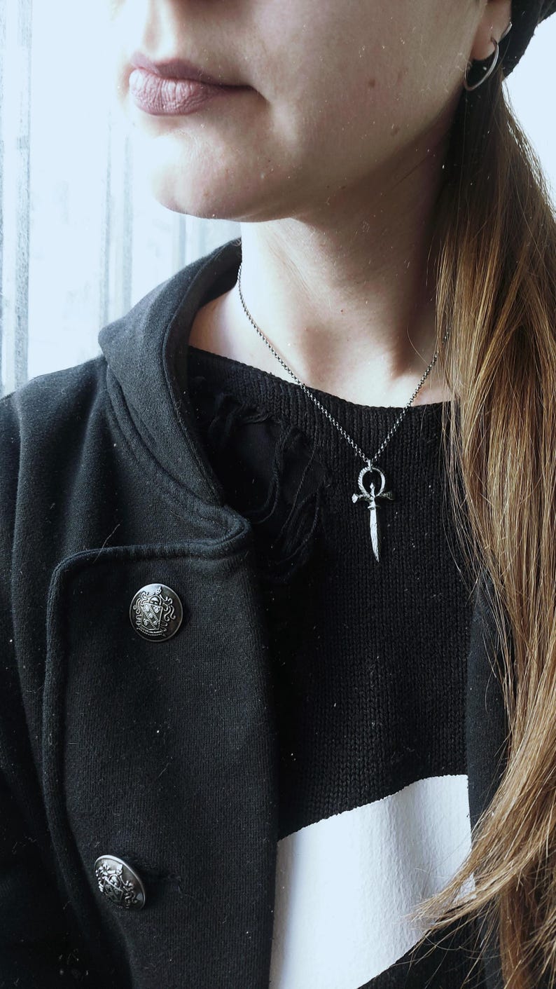 May include: A person wearing a black hooded jacket with a silver dagger pendant necklace. The pendant is a stylized dagger with a circle at the top. The person is wearing a black and white striped shirt underneath the jacket.