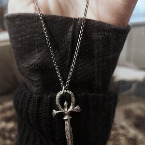 May include: A silver chain necklace with a silver pendant shaped like an ankh with a dagger in the center.