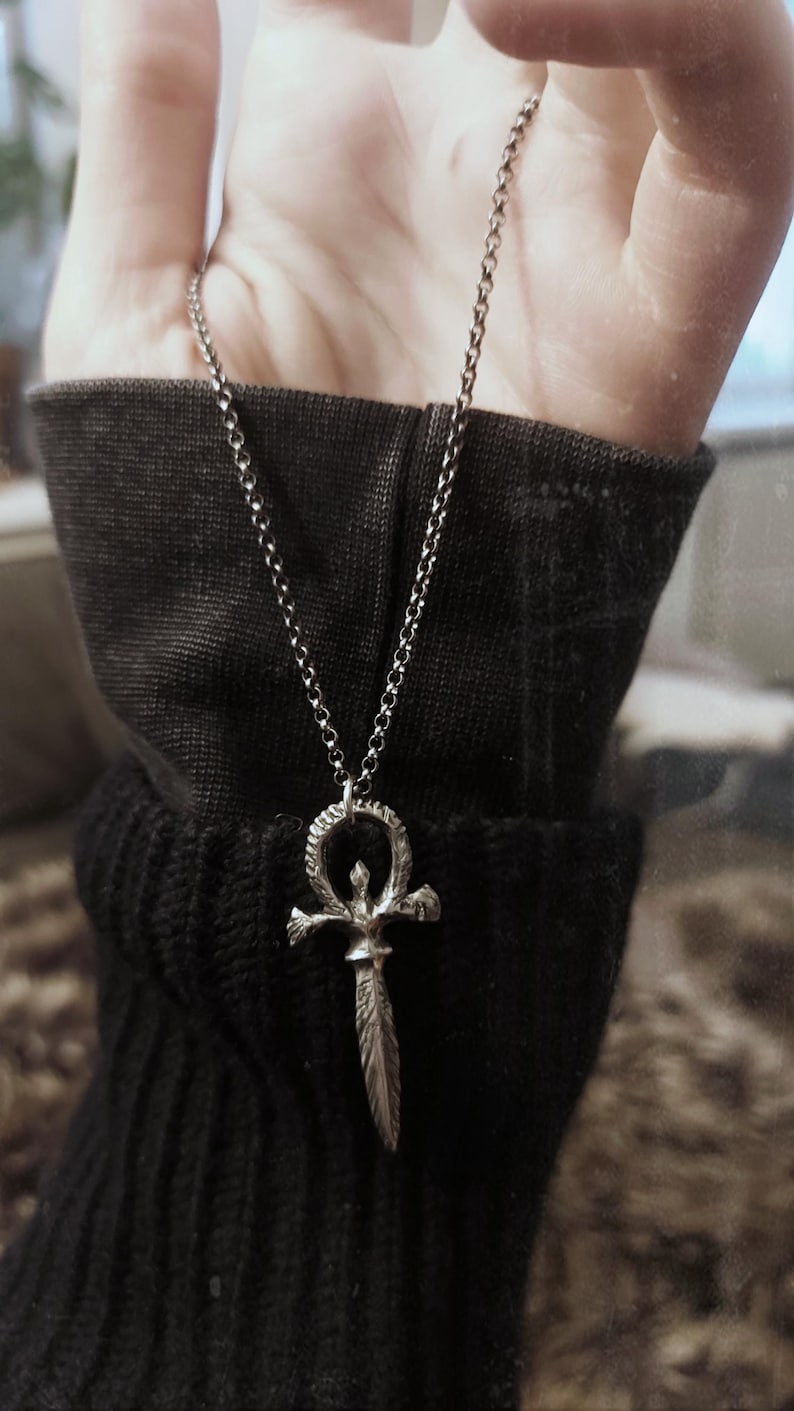 May include: A silver chain necklace with a silver pendant shaped like an ankh with a dagger in the center.