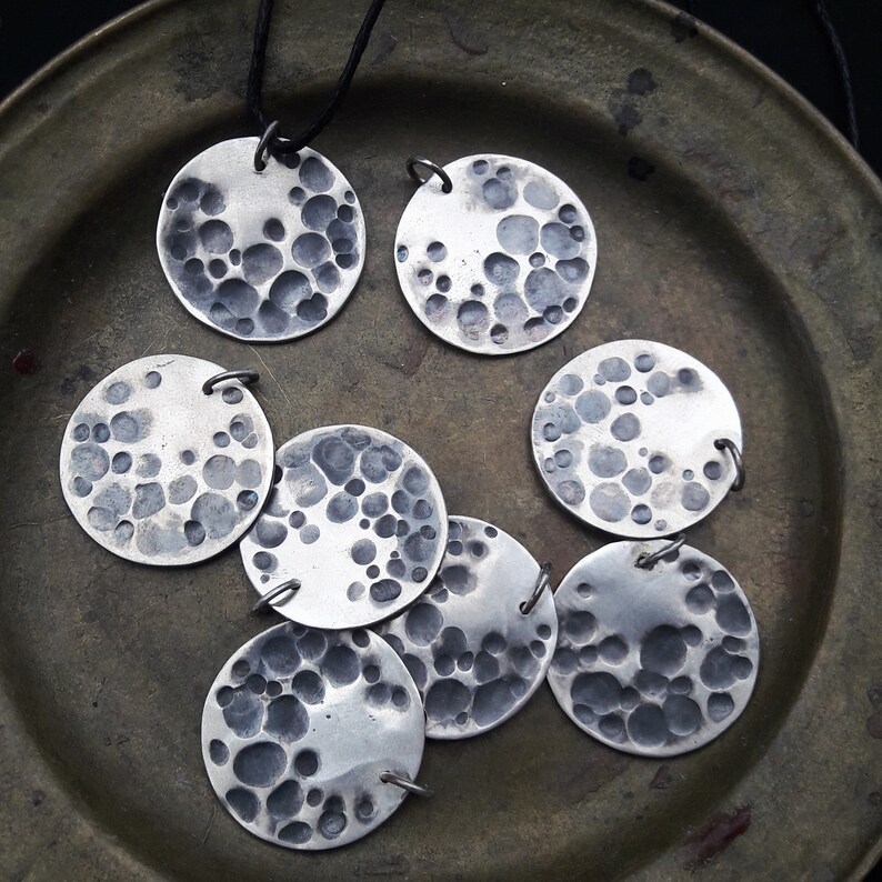 May include: A set of seven silver pendants, each with a circular shape and a textured surface resembling the moon's surface. The pendants are arranged on a dark brown surface.