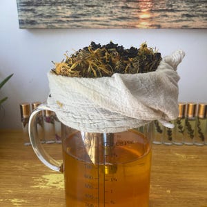 May include: A clear glass measuring pitcher filled with golden liquid, featuring markings for milliliters and US pints. A white cloth filter sits on top, containing a mound of dried yellow and dark brown botanical herbs and plant matter.