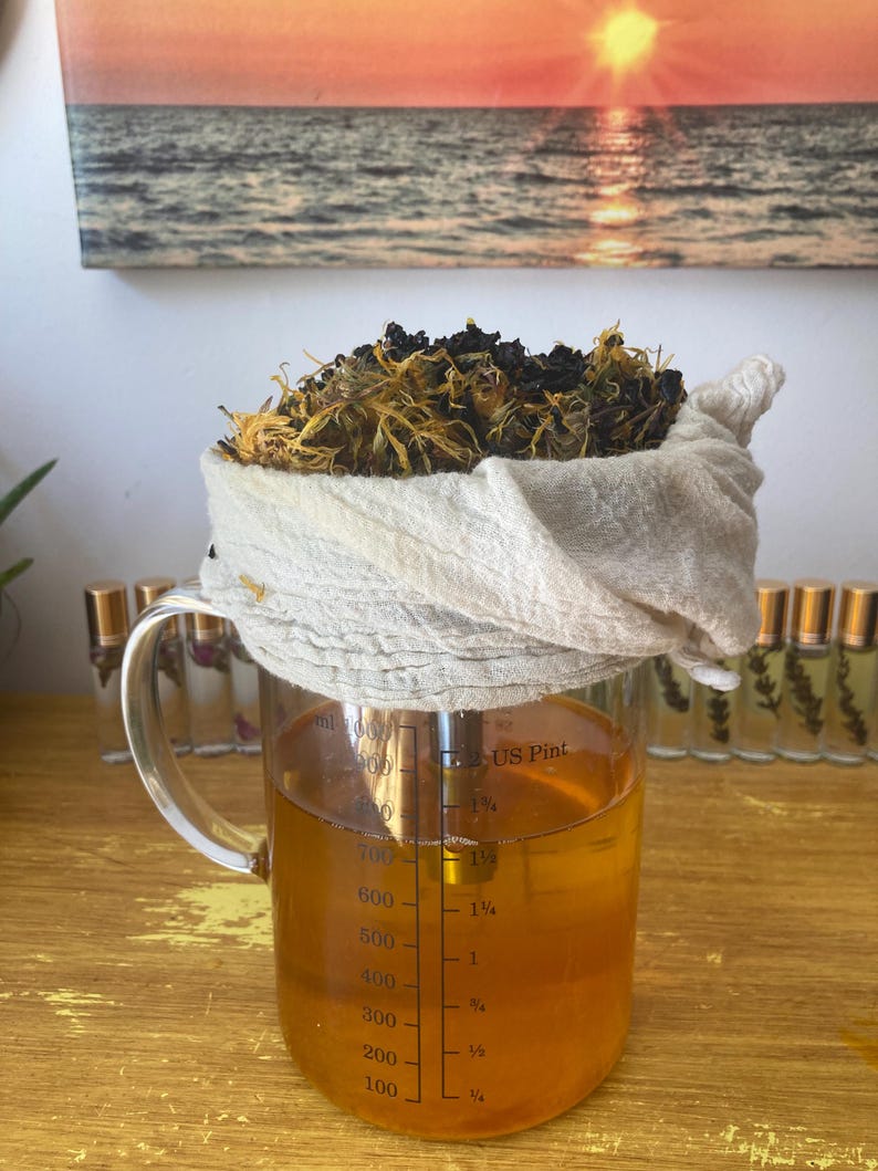 May include: A clear glass measuring pitcher filled with golden liquid, featuring markings for milliliters and US pints. A white cloth filter sits on top, containing a mound of dried yellow and dark brown botanical herbs and plant matter.