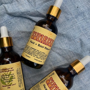 May include: Three amber glass bottles of Sunrise Face + Body Serum by Hippie Twig. Each bottle features a gold dropper cap and a cream-colored label with red text, listing ingredients like cranberry seed oil, marula oil, and elderberry extract.