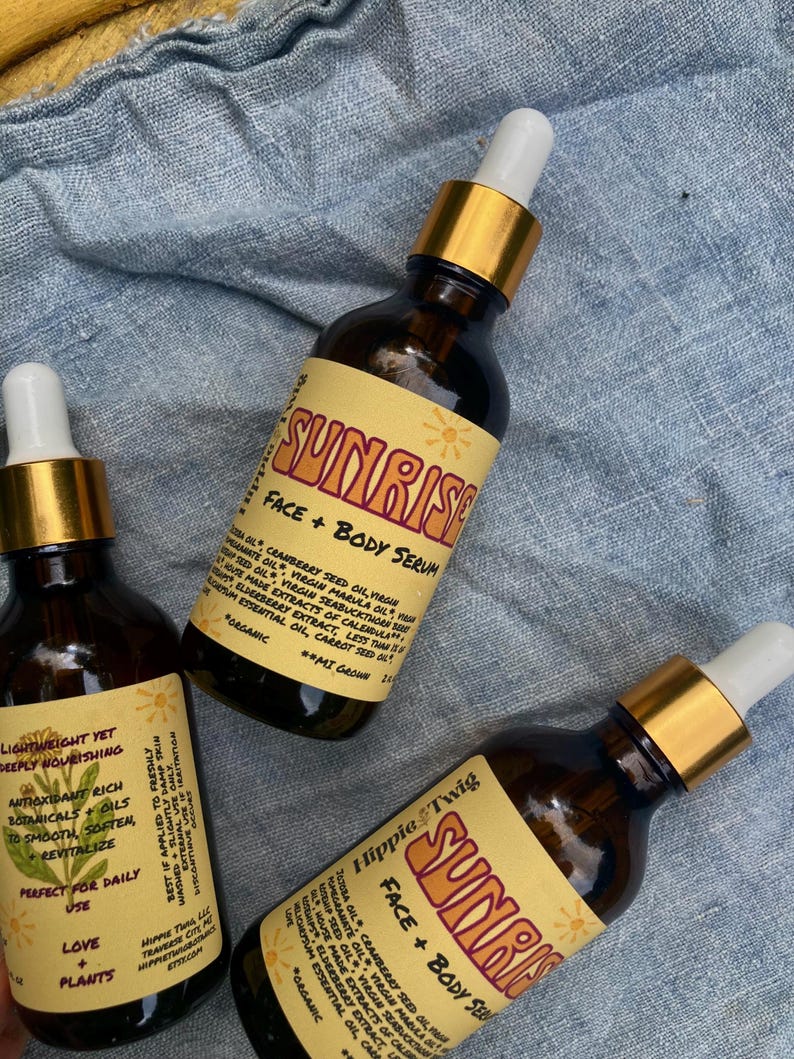 May include: Three amber glass bottles of Sunrise Face + Body Serum by Hippie Twig. Each bottle features a gold dropper cap and a cream-colored label with red text, listing ingredients like cranberry seed oil, marula oil, and elderberry extract.
