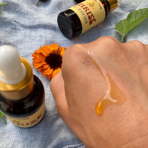 May include: Two amber glass dropper bottles labeled Sunrise Repair Serum with gold caps and white droppers. A drop of golden liquid serum is visible on skin. Nearby are a bright orange calendula flower, dried rose hips, and a green leaf.