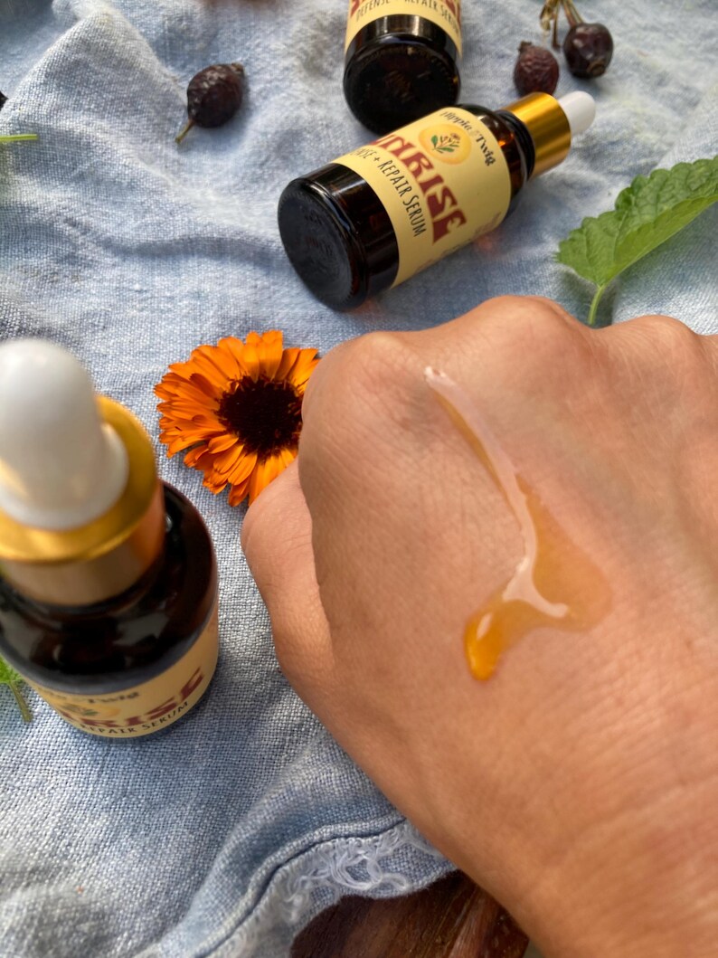 May include: Two amber glass dropper bottles labeled Sunrise Repair Serum with gold caps and white droppers. A drop of golden liquid serum is visible on skin. Nearby are a bright orange calendula flower, dried rose hips, and a green leaf.