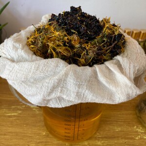 May include: A clear glass measuring pitcher filled with a golden liquid, topped with a white cheesecloth containing a mound of dark brown and yellow dried botanical matter. The pitcher features black measurement markings up to 800 milliliters.