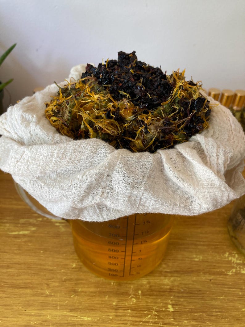 May include: A clear glass measuring pitcher filled with a golden liquid, topped with a white cheesecloth containing a mound of dark brown and yellow dried botanical matter. The pitcher features black measurement markings up to 800 milliliters.