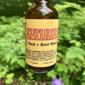 May include: A brown glass bottle with a gold cap containing face and body serum. The yellow label features the text SUNRISE, FACE + BODY SERUM, and Hippie & Twig, along with a list of organic ingredients including jojoba, cranberry, and marula oil.