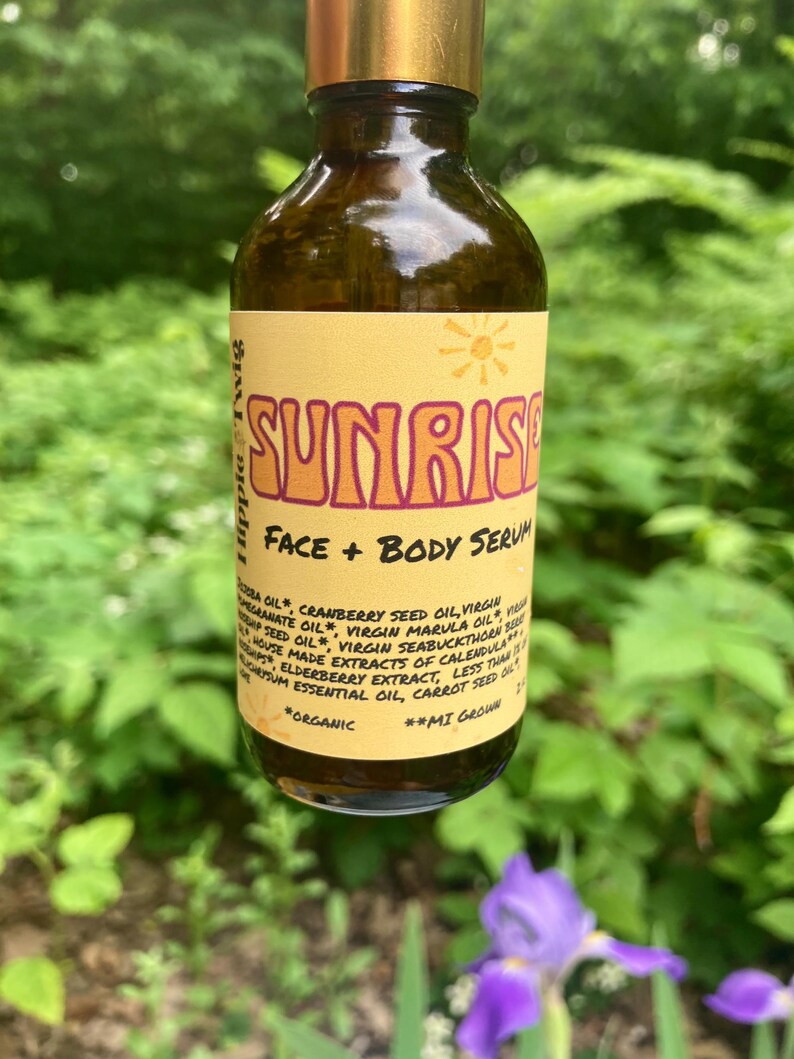 May include: A brown glass bottle with a gold cap containing face and body serum. The yellow label features the text SUNRISE, FACE + BODY SERUM, and Hippie & Twig, along with a list of organic ingredients including jojoba, cranberry, and marula oil.