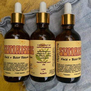 May include: Three amber glass dropper bottles of Sunrise Face + Body Serum by Hippie Twig. Each bottle features a yellow label with brown text, a botanical illustration, and a list of organic ingredients including jojoba, cranberry, and marula oil.