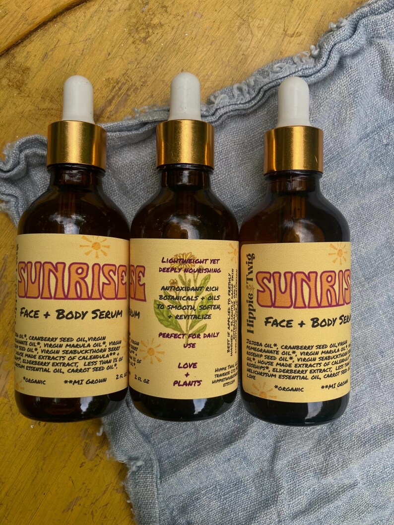 May include: Three amber glass dropper bottles of Sunrise Face + Body Serum by Hippie Twig. Each bottle features a yellow label with brown text, a botanical illustration, and a list of organic ingredients including jojoba, cranberry, and marula oil.