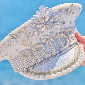 May include: White bridal cap adorned with sparkling rhinestones, pearls, and sequins. The word "BRIDE" is spelled out in large, shimmering letters across the front. A decorative accessory for a wedding.