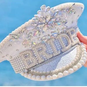 May include: A white captain-style hat featuring the word BRIDE in large pearl-encrusted letters across the front. The hat is heavily decorated with iridescent crystals, silver rhinestones, and a border of white pearls along the brim and crown.