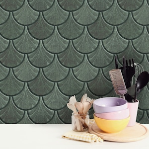 May include: A set of kitchen utensils, including a grater, a whisk, and a spatula, are arranged in a glass jar. The utensils are placed on a white countertop in front of a green and gray patterned wall. There are also three bowls, one purple, one pink, and one yellow, stacked on top of each other. The bowls are on a wooden cutting board.