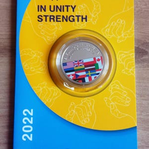 May include: A silver coin with the text "В ЄДНОСТІ – СИЛА" and "IN UNITY STRENGTH" in Ukrainian and English. The coin features a design of hands clasped together and a circle of flags representing different countries. The coin is displayed on a blue and yellow card with the text "МОНЕТИ УКРАЇНИ" and "COINS OF UKRAINE" in Ukrainian and English. The card also includes the year "2022", the text "Національний банк України" and "National Bank of Ukraine" in Ukrainian and English.