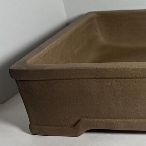 May include: A rectangular, matte brown ceramic bonsai pot with a wide, flat rim and a recessed base featuring small feet at each corner. The interior is deep and smooth, showing a consistent earthy tone throughout the entire exterior surface.