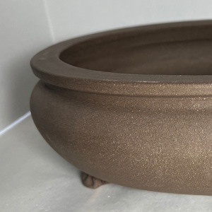 May include: A brown oval ceramic planter with three small feet. The planter has a smooth, slightly textured surface.