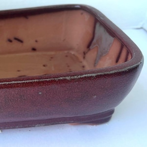 May include: A square, ceramic planter with a burgundy exterior and a lighter brown interior. The planter has a slightly textured surface and sits on a small, rectangular base. The rim has a white accent.