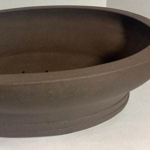 May include: A dark brown, oval-shaped ceramic planter with a smooth, matte finish. The vessel features a wide, open top and a slightly raised, rounded base. The interior is deep and hollow, showing a simple, unadorned surface with drainage holes.