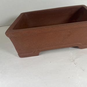 May include: A rectangular, terracotta-colored bonsai pot. The pot has a slightly textured surface and a simple, classic design. It features a raised rim and small feet for stability. Ideal for small trees.
