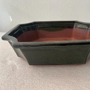 May include: A dark green ceramic bonsai pot with a rectangular shape and flared sides. The interior has a reddish-brown color. The pot has a glossy finish and is designed for planting small trees or plants.