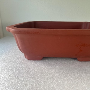 May include: A terracotta-colored rectangular bonsai pot with a slightly fluted design. The pot has four small feet and a wide, open top. The overall shape is square with rounded corners, suitable for small trees.