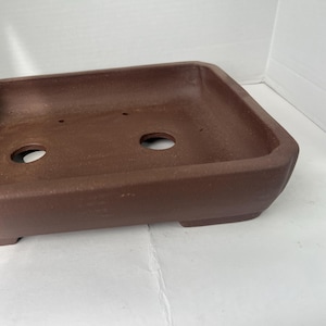 May include: A rectangular, brown bonsai pot with a low profile. The pot has a slightly textured surface and two drainage holes. The pot is made of a durable material and is designed for growing bonsai trees.