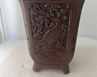 May include: A brown ceramic planter with a detailed relief carving of a bird and foliage. The rectangular pot has a flared top, small feet, and drainage holes. A decorative piece for plants.
