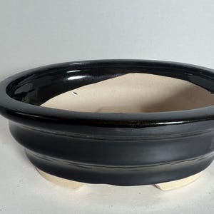 May include: A black oval ceramic planter with a glazed finish and a slightly raised rim. The planter has three small feet on the bottom.