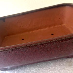 May include: A rectangular bonsai pot with a reddish-brown interior and a dark burgundy exterior. The pot has a slightly textured surface and two drainage holes. The edges are rounded, and the pot sits on a low base.