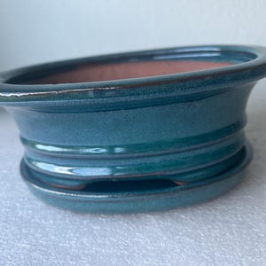 May include: A shallow, oval-shaped ceramic bonsai pot with a deep teal, glossy glaze. The exterior features horizontal ridges along the sides, and the interior reveals a natural terracotta-colored clay finish. It sits on a matching teal saucer.