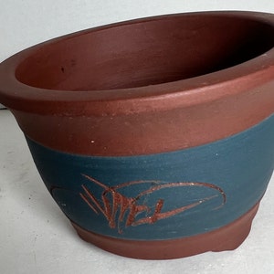 May include: A terracotta bonsai pot with a dark blue band and a brown design. The pot has a rounded shape and a wide opening.