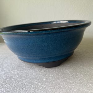 May include: A round, deep ceramic bonsai pot featuring a vibrant, glossy blue glaze on the exterior and a dark, matte finish on the interior and base. The bowl has a wide, slightly flared rim and a sturdy, elevated footed bottom design.