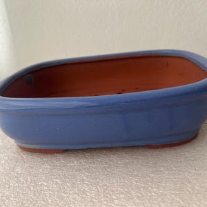 May include: A rectangular bonsai pot with rounded corners, featuring a smooth, glossy periwinkle blue glazed exterior and a contrasting unglazed terracotta interior. The base is elevated with small feet, and drainage holes are visible inside.