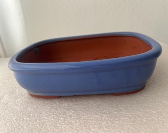 May include: A rectangular bonsai pot with rounded corners, featuring a smooth, glossy periwinkle blue glazed exterior and a contrasting unglazed terracotta interior. The base is elevated with small feet, and drainage holes are visible inside.