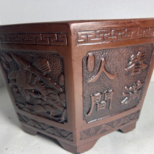 May include: A brown terracotta pot with a square shape and intricate carvings. The pot features a design of a bird and a tree on one side and Chinese characters on the other side. The characters read "人寿" which translates to "long life".