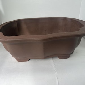 May include: A brown, octagonal bonsai pot with a scalloped rim. The pot has a low profile with a slightly textured surface and sits on four small feet. The overall shape is symmetrical and designed for displaying bonsai trees.