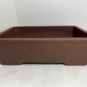 May include: A rectangular brown bonsai pot with a slightly tapered design. The pot has a simple, minimalist aesthetic, suitable for showcasing bonsai trees. The pot is made of a durable material and features a smooth, matte finish.