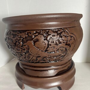 May include: A brown ceramic planter with a raised design of birds and flowers. The planter has a wide base with three legs.