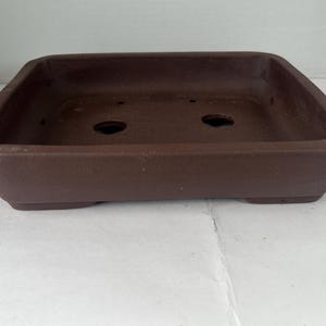 May include: A square, brown bonsai pot with a rectangular shape. The pot has a low profile with a flat base and slightly angled sides. Two drainage holes are visible in the bottom of the pot. The pot is made of a matte, earthy material.