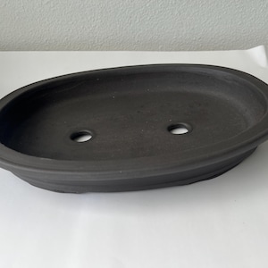 May include: A dark gray, oval-shaped bonsai pot with two drainage holes. The pot has a slightly raised rim and a smooth, matte finish. The pot is designed for planting and cultivating bonsai trees.