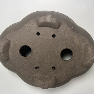 May include: A brown, oval-shaped bonsai pot with a textured surface. The pot has three feet and several drainage holes. The pot is approximately 12 inches long and 9 inches wide.