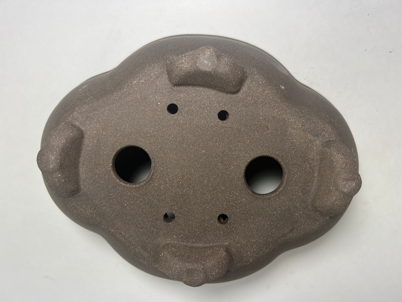 May include: A brown, oval-shaped bonsai pot with a textured surface. The pot has three feet and several drainage holes. The pot is approximately 12 inches long and 9 inches wide.